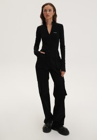 Juodas ilgomis rankovėmis viršus su užtrauktuku apykakle, derinamas su juodais cargo kelnėmis, turinčiomis daugybę kišenių ir prisiūtą fasoną, vilkimas su masyviais juodais batais.