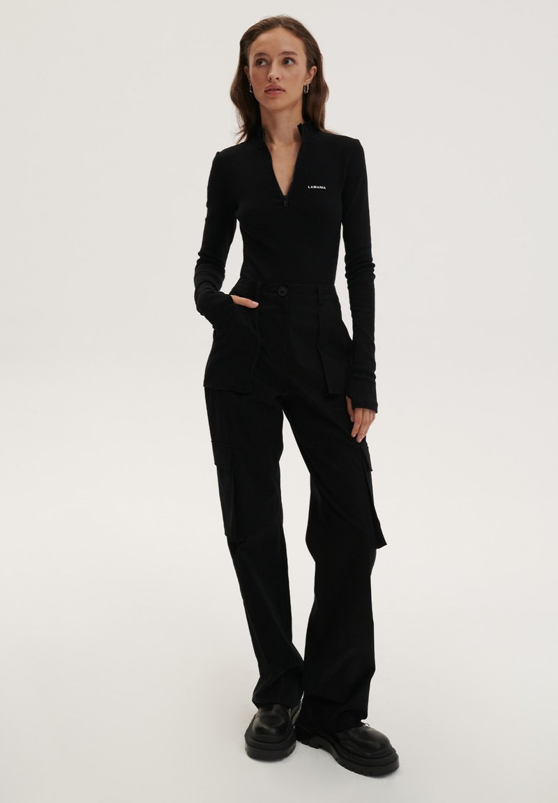 Juodas ilgomis rankovėmis viršus su užtrauktuku apykakle, derinamas su juodais cargo kelnėmis, turinčiomis daugybę kišenių ir prisiūtą fasoną, vilkimas su masyviais juodais batais.