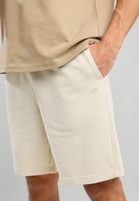 Vaaleanbeige puuvillapaita, jossa on pyöristetty helma, yhdistettynä kermaisiin shortseihin, joissa on sivutaskut ja pieni logo vasemmassa reidessä.