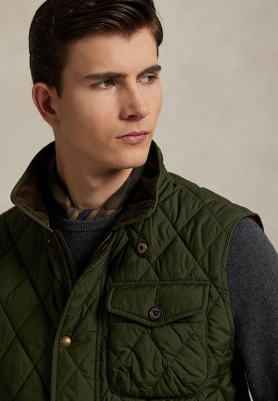 Joven con un chaleco acolchado verde oscuro sobre un suéter gris, mirando hacia su izquierda contra un fondo neutro y sencillo.