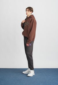 Ruskea huppari, jossa on etutasku, yhdistettynä tummanharmaisiin collegehousuihin, joissa on purppura logo, ja valkoisiin urheilujalkineisiin.