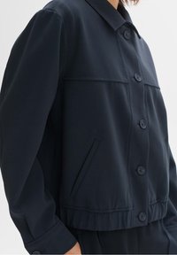 Granatowa krótka kurtka wykonana z gładkiej tkaniny, posiadająca zapięcie na guziki z przodu, dwie kieszenie na piersi oraz usztywniony kołnierz z lekko marszczonym dołem.