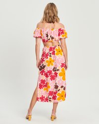 Off-the-shoulder komplet s kratkim topom in maksi krilom v roza barvi z velikimi cvetličnimi vzorci v rdeči, rumeni in rjavi. Elastičen pas in bočni razporek.