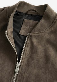 Chaqueta bomber de ante marrón con un zipper plateado y un cuello acanalado. El interior tiene un forro negro y una etiqueta de tela cosida por dentro.