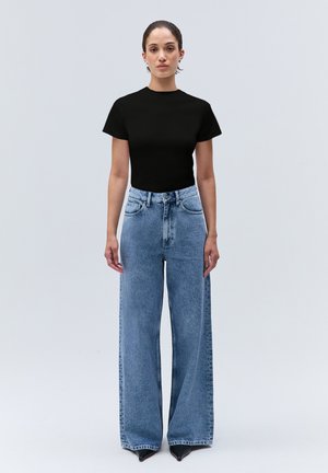 Femme debout portant un haut noir à manches courtes et un jean bleu taille haute à jambes larges avec des chaussures noires pointues sur un fond uni.