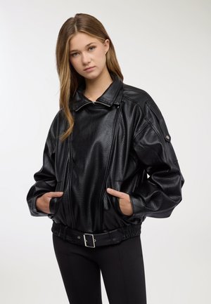 Giovane donna con capelli lunghi che indossa una giacca di pelle nera testurizzata oversize, con le mani in tasca, in piedi contro uno sfondo chiaro e uniforme.