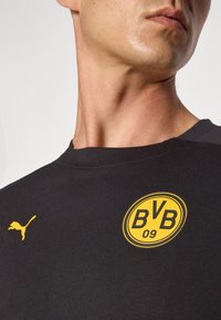 Mustan lyhythihaisen t-paidan, jossa on keltainen BVB 09 -logo ja pieni keltainen Puma-embleemi vasemmalla olkapäällä, valmistettu sileästä puuvillasekoitekankaasta.