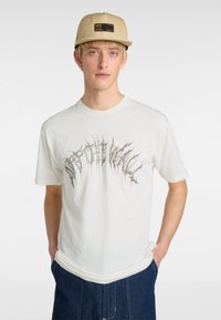 Hvid bomulds t-shirt med et grafisk design med skarpe, spidse bogstaver. Modellen bærer en beige kasket med et sort logo og sidelommer.
