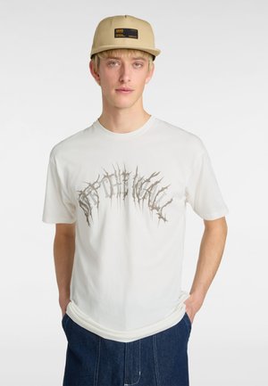 White cotton t-shirt featuring a graphic design with sharp, spiky text. The model wears a beige cap with a black logo and side pockets.