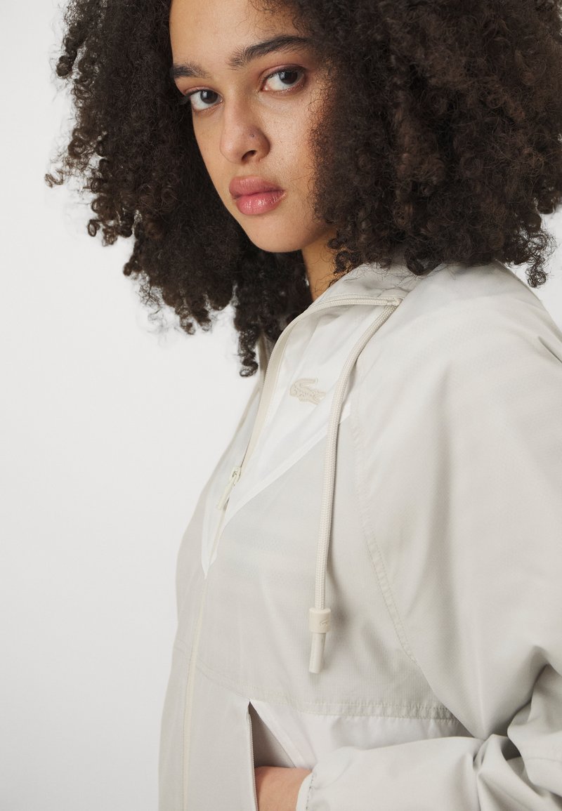 Lichtbeige windbreaker met een rits voorkant, een capuchon met trekkoord en zijzakken. De stof heeft een gladde textuur met subtiele patronen.