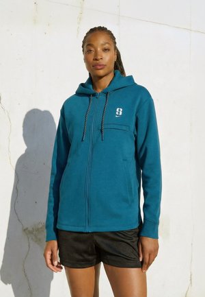 Kvinne iført teal hettejakke med glidelås og hette, samt svarte shorts, står mot en hvit vegg i sollys.