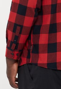 Camisa de franela a rayas en rojo y negro, con mangas largas, puños abotonados y un dobladillo curvado. La textura de la tela parece suave y ligeramente cepillada.
