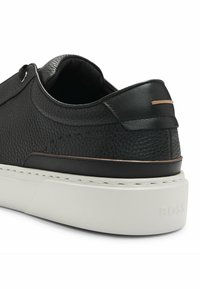 Zapatilla de cuero negro con un acabado texturizado, suela de goma blanca y un sutil acento en bronce en el talón. Presenta detalles de costura mínimos.