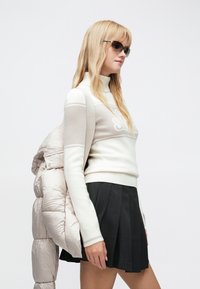 Lyshåret kvinde iført solbriller, hvid rullekrave-trøje, sort plisseret nederdel, holder en lysebeige dunjakke over skulderen.