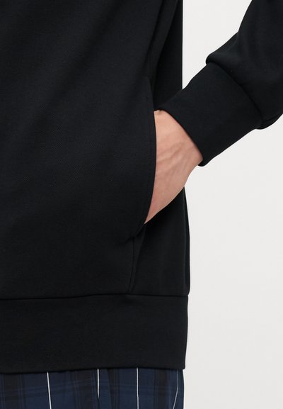Black, textured sweatshirt featuring a kangaroo pocket and ribbed cuffs, worn over navy plaid pants with white stripes.