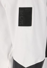 Vitt jacka med svarta detaljer, inklusive en texturerad logotypmärket som säger "SLOPE STYLE". Tyget ser slätt ut med en modern design.