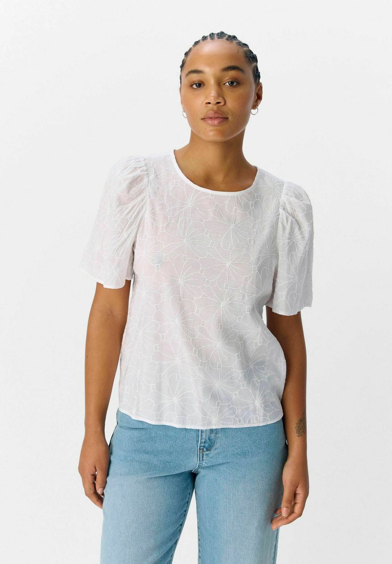 Witte blouse met korte mouwen, voorzien van een gestructureerd bloemenpatroon, ronde halslijn en opgeblazen mouwen, gedragen over lichtblauwe denim jeans.