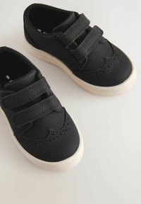Schwarze Kinderschuhe mit zwei Klettverschlüssen, glattem synthetischen Material, Brogue-Dekor und einer kontrastierenden weißen Gummisohle.
