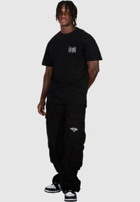 Sort kortærmet t-shirt med logo på fronten, parret med sorte cargo-bukser med sidelommer og hvide grafiske detaljer; sort og hvide sneakers.