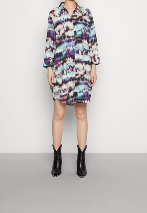 Personne debout contre un mur uni, portant une robe chemise tie and dye multicolore à manches longues, longueur genou, et des bottines noires.