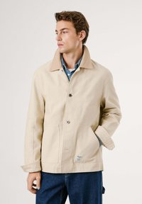 Joven vestido con una chaqueta beige sobre una camisa de mezclilla azul y jeans azules, de pie con una mano en el bolsillo, mirando a la izquierda sobre un fondo liso.