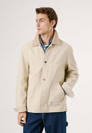 Giovane uomo che indossa una giacca beige sopra una camicia di jeans blu e jeans blu, in piedi con una mano in tasca, che guarda a sinistra su uno sfondo semplice.