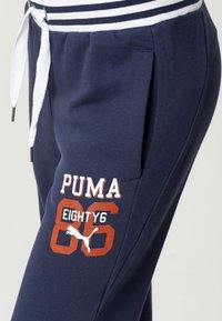 Navy jogger pants with white striped elastic waistband, featuring a drawstring, side pockets, and bold "PUMA 86 EIGHTY6" graphic in red and white.