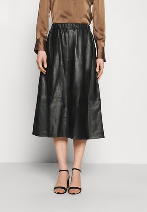 Femme portant une jupe midi en similicuir noir avec une ceinture élastique, associée à un chemisier en satin marron et des escarpins noirs à bride à la cheville.