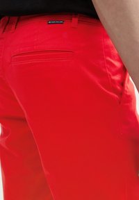Pantalones de algodón rojos con un corte ajustado, que cuentan con un bolsillo trasero y una etiqueta de logo. La textura suave y el color brillante dominan el diseño.