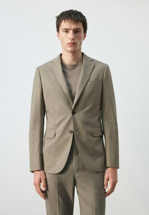 Joven con chaqueta de traje beige y pantalones a juego sobre una camiseta de cuello redondo taupe, de pie contra un fondo blanco liso.