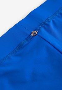 Close-up af blåt stof med forstærket syning og et lille rundt guldfarvet metallogo fastgjort nær en søm.