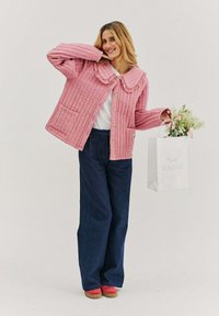 Růžová prošívaná bunda s velkým límcem, knoflíkovým zapínáním a dvěma předními kapsami. V kombinaci s tmavě modrými kalhotami a červenými botami, drží bílou tašku s květinami.