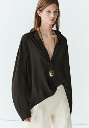 Junge Frau trägt eine lockere schwarze Bluse, weiße Hose und eine lange Halskette mit großem goldenen Anhänger und schwarzer Quaste.