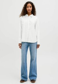 Witte katoenen overhemd met lange mouwen en een kraag, gecombineerd met lichtblauwe uitlopende spijkerbroek en donkerbruine laarzen. Eenvoudige, casual outfit.