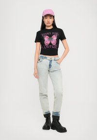 Svart kortermet t-skjorte med rosa sommerfugl-grafikk og teksten "Versace Jeans Couture". Lyseblå denimjeans med oppbrettede hem og svarte støvler. Rosa caps.