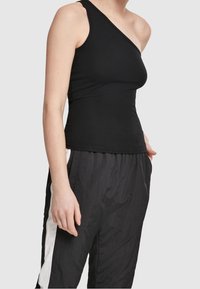 Personne portant un top noir cintré une épaule et un pantalon noir ample avec des bandes blanches sur les côtés, debout avec une main dans la poche.