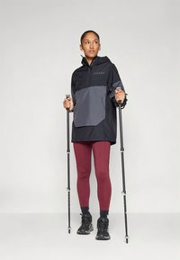Woman wearing black and gray windbreaker and maroon leggings holding two trekking poles, standing on a plain light background.