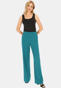 Czarny top bez rękawów w połączeniu z turkusowymi spodniami o szerokich nogawkach, charakteryzującymi się gładką teksturą i elastycznym pasem. Model nosi czarne sandały na obcasie.