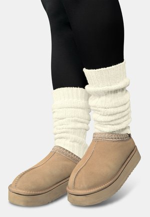 Slip-on Schuhe aus hellbraunem Wildleder mit strukturierten Sohlen, verziert mit gewebten Details oben. Cremafarbene, gestrickte Stulpen werden über schwarzen Leggings getragen.