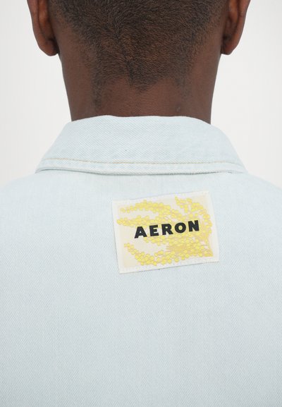 Casaco de ganga azul claro com uma etiqueta que apresenta gráficos botânicos amarelos e o texto "AERON" em preto, visível na parte de trás do colarinho.