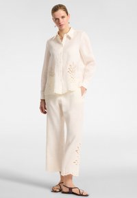 Blusa in lino leggero color crema con dettagli ricamati all'orlo e alle maniche, abbinata a pantaloni a gamba larga coordinati e sandali.