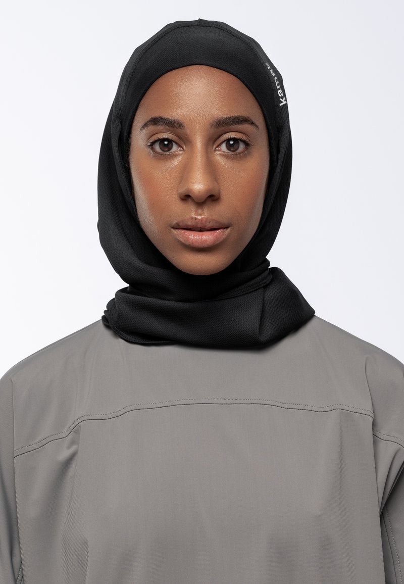 Donna con un hijab nero e una maglietta grigia, che guarda dritto davanti a sé con espressione neutra su uno sfondo bianco uniforme.