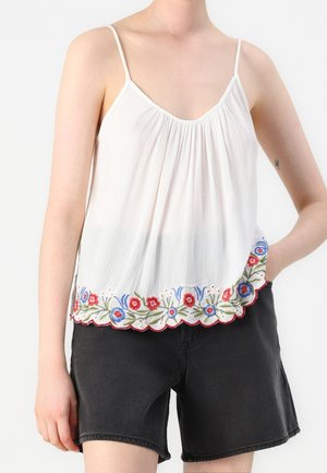Vrouw draagt een wit mouwloos topje met bloemenborduursel aan de zoom en zwarte spijkershorts tegen een effen achtergrond.
