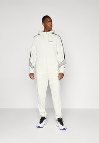 Sudadera blanca con cremallera y acentos en negro y gris, combinada con joggers blancos a juego. Logo de la marca en el pecho, tejido texturizado, corte casual.