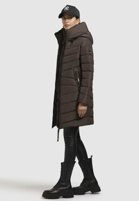 Giacca a vento marrone con colletto alto, design a righe orizzontali, tasche laterali e chiusura con zip, abbinata a leggings in faux pelle neri e stivali con lacci.