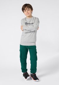 Ragazzo in piedi con le braccia incrociate, indossa una felpa grigia della Champion, pantaloni cargo verdi e scarpe da ginnastica nere con suole bianche.