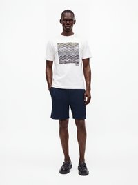 Homme portant un t-shirt blanc à manches courtes avec un motif ondulé et un short bleu marine, debout les mains dans les poches sur fond blanc.