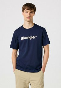 Mørkeblå t-shirt lavet af bomuld, med et stort hvidt "Wrangler" logo på tværs af brystet. Enkel rund hals og korte ærmer.