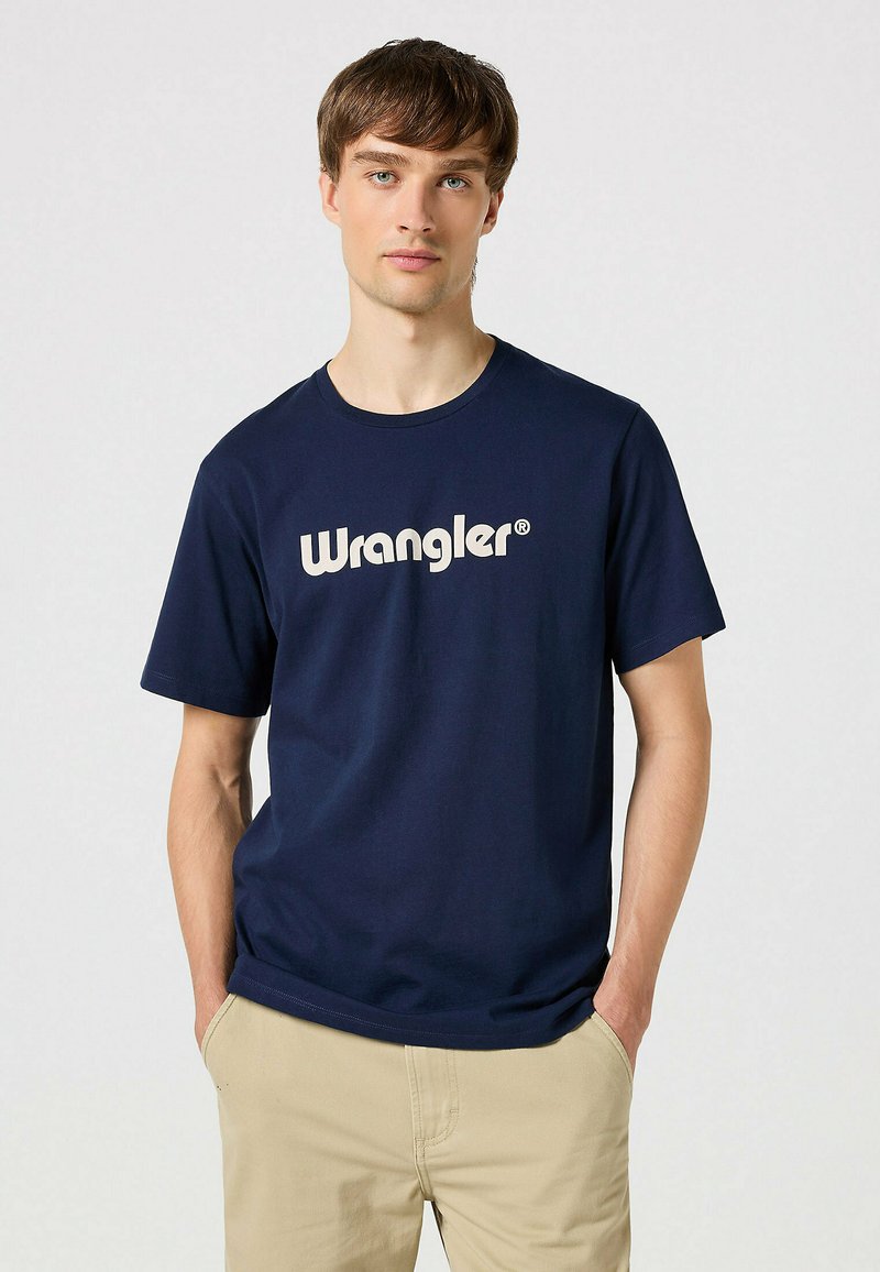 Mørkeblå t-shirt lavet af bomuld, med et stort hvidt "Wrangler" logo på tværs af brystet. Enkel rund hals og korte ærmer.