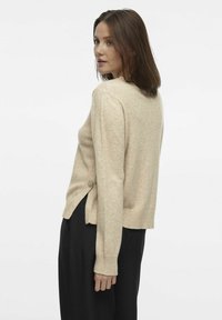 Beige kootud sviiter, millel on ümar kaelus, soonikkoes varrukad ja dekoratiivne nööp külgmise ääre peal. Kanna mustade pükstega.